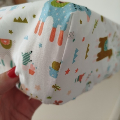 Máscara de tecido branco com padrão colorido de animais e estrelas segurada por uma mão.