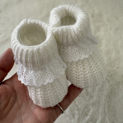 Botins de bebé em malha branca com renda na gola, segurados numa mão.