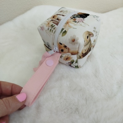 Estojo quadrado com padrão floral e animais, fecho branco e alça rosa sobre superfície branca macia