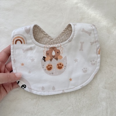 Babete de bebé bege e branco com padrão de dinossauro, ossos e arco-íris