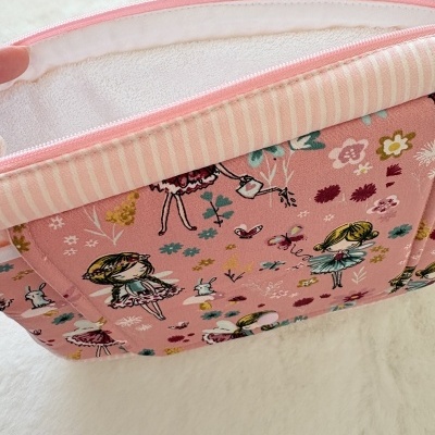 Mala de tecido rosa com padrão de bonecas e flores coloridas, fecho rosa, interior branco macio e borda rosa às riscas