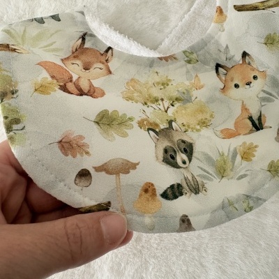 Babete com padrão de animais e folhas em tecido macio branco.
