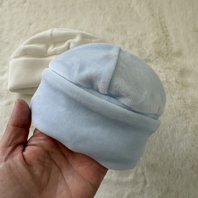Dois gorros de bebé azuis e branco em tecido de veludo macio sobre superfície bege.