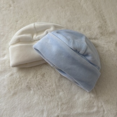Dois gorros de bebé, branco e azul claro, sobre superfície felpuda bege