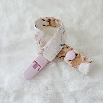 Prendedor de chupeta estampado com clip e botão rosa numa superfície branca macia.