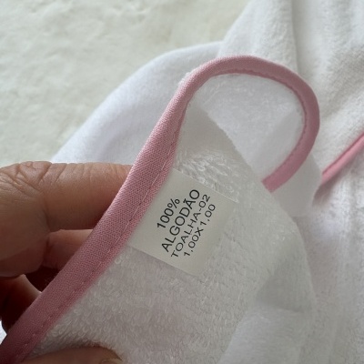 Toalha branca de algodão com rebordo rosa claro segurada por uma mão e detalhe na etiqueta