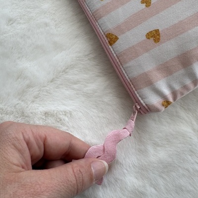 Bolsa ou estojo de tecido rosa com corações amarelos e riscas brancas, fecho de correr rosa, sobre superfície branca felpuda, mão segurando puxador