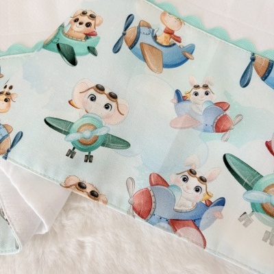 Tecido infantil com animais em aviões coloridos e borda verde sobre pano branco.