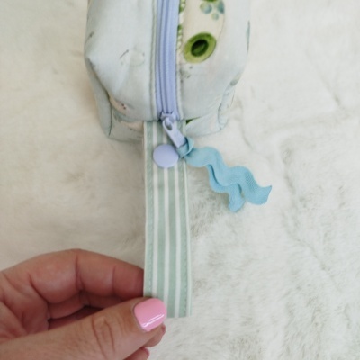 Estojo branco floral com fecho de correr lilás e puxadores coloridos, mão segurando fita listrada verde e branca