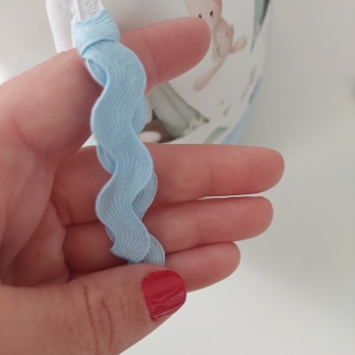 Fita azul clara ondulada entre dedos com unhas vermelhas e urso de peluche ao fundo