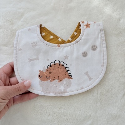 Babete dupla-face para bebé com padrão de dinossauro e estrelas sobre tecido branco felpudo.