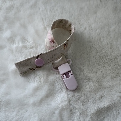 Alça para chupeta em tecido estampado com fecho de plástico rosa sobre superfície macia creme