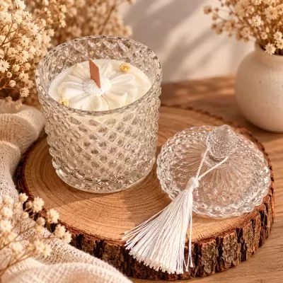 Vela decorativa branca em vaso de vidro facetado com tampa ao lado e flores secas