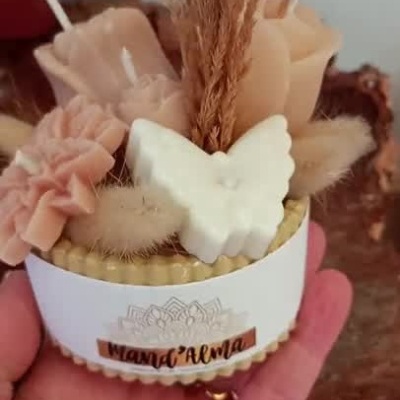 Arranjo decorativo em cera com velas florais, borboleta branca e elementos secos na mão.