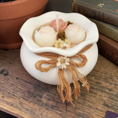 Vela decorativa branca com flores e laço de cordão, sobre mesa de madeira junto a suculenta e livros.
