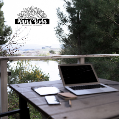 Computador portátil e acessórios numa mesa de madeira numa varanda com vista para o lago e árvores.