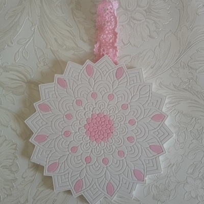 Enfeite branco e rosa em forma de mandala com fita rosa pendurado sobre tecido branco com padrão floral