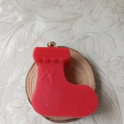 Botão decorativo vermelho em forma de bota de Natal sobre disco de madeira em fundo branco texturizado.