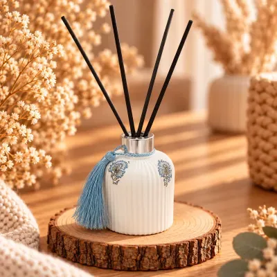 Difusor de aromas branco com detalhes florais e franja azul numa base de madeira com flores secas em ambiente acolhedor.