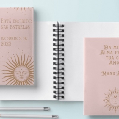 Três cadernos espiral rosa com desenhos dourados do sol, estrelas e lua em fundo azul claro, acompanhados de dois lápis e um pires.