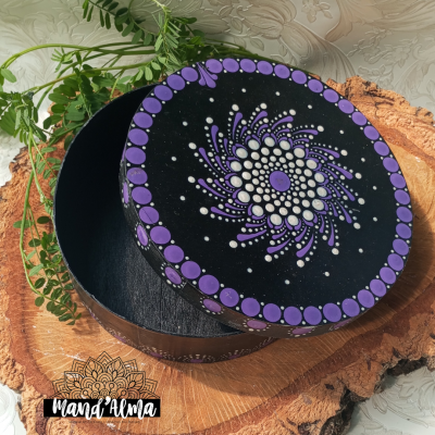 Caixa redonda preta com padrões de pontos lilás e branco sobre tampa, em superfície de madeira.