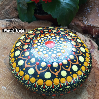 Pedra pintada com padrão mandala colorido sobre madeira, com planta e parede de pedra ao fundo