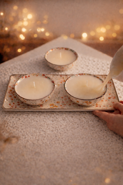 Velas em tigelas de cerâmica floral sobre tabuleiro com padrão semelhante sobre uma mesa texturizada bege.