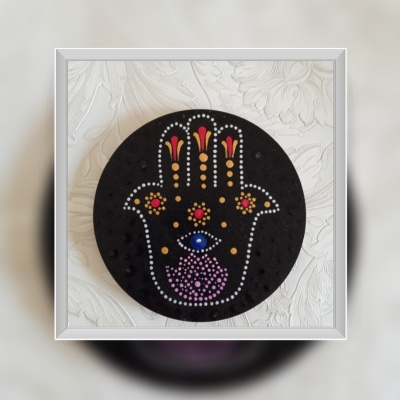 Objeto decorativo redondo preto com padrão colorido de mão de Hamsá sobre superfície branca texturizada