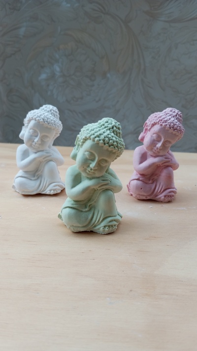 Três estatuetas de Buda em cerâmica, cores branco, verde e rosa, sobre superfície de madeira