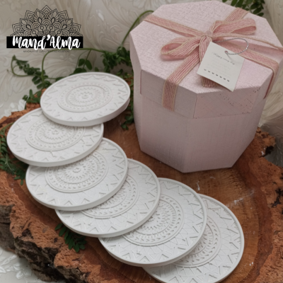 Bases redondas brancas com padrões geométricos em madeira rústica, ao lado de caixa rosa com laço e etiqueta.