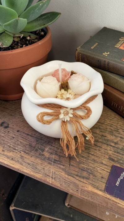 Vela decorativa branca com flores e laço de cordão, sobre mesa de madeira junto a suculenta e livros.