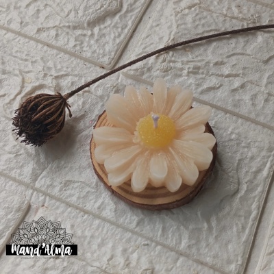 Vela em forma de flor branca com centro amarelo sobre base de madeira com flor seca ao lado em azulejos brancos rugosos