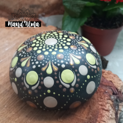 Pedra pintada com padrões de pontos coloridos sobre madeira, ao lado de planta em vaso.