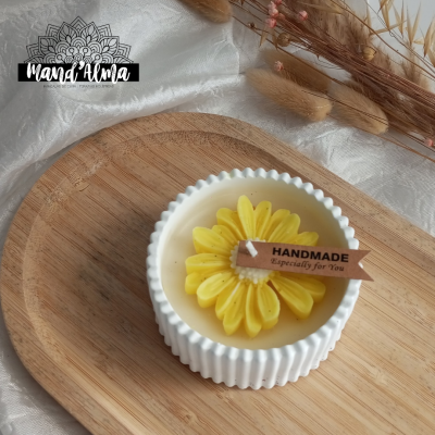 Vela artesanal amarela em forma de flor dentro de recipiente branco sobre tábua de madeira