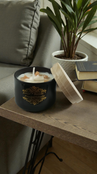 Vela branca em recipiente preto com tampa de madeira clara sobre mesa de madeira próxima a sofá cinzento e planta em vaso branco.