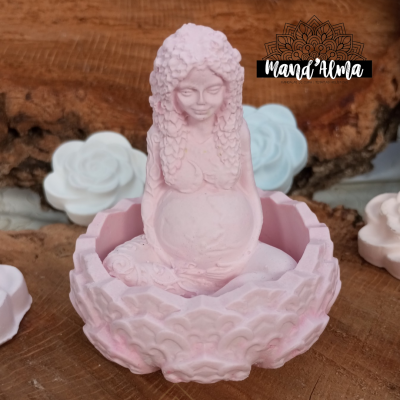 Incensário cor-de-rosa em forma de mulher grávida em base decorativa floral, sobre madeira, com flores brancas ao fundo e logo 'Mana Alma'.