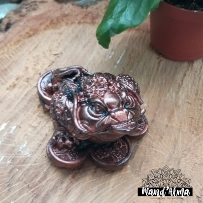 Estátua de sapo decorativa em metal bronze com olhos verdes sobre superfície de madeira com planta ao lado