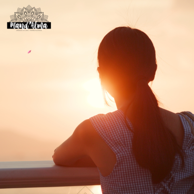 Jovem de cabelo castanho escuro e vestido de xadrez azul claro e branco a olhar para o pôr do sol encostada a um corrimão branco