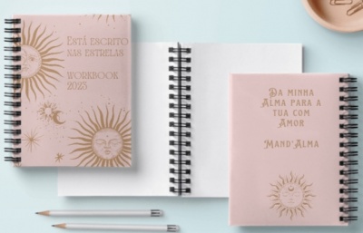 Três cadernos espiral rosa com desenhos dourados do sol, estrelas e lua em fundo azul claro, acompanhados de dois lápis e um pires.