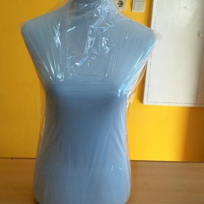 Manequim feminino cinza claro com plástico transparente numa mesa contra parede amarela