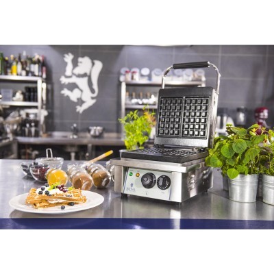 Máquina de waffles em metal inox com painéis pretos numa cozinha profissional com waffles e plantas em vasos.