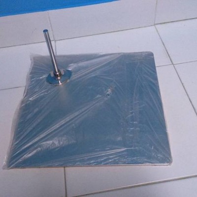 Base metálica quadrada com suporte cilíndrico coberta por película plástica sobre chão de azulejos brancos.