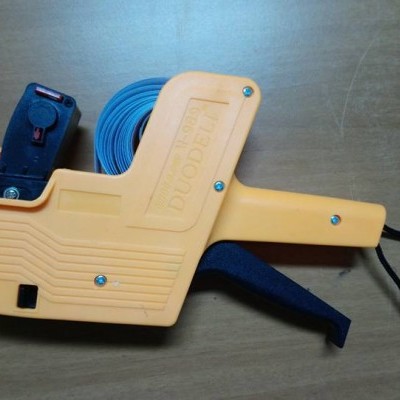 Etiquetadora manual plástica amarela com fita azul e alça preta sobre madeira