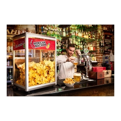 Expositor transparente cheio de nachos amarelos com placa 'Mexican Nachos' em balcão de bar onde homem serve cerveja