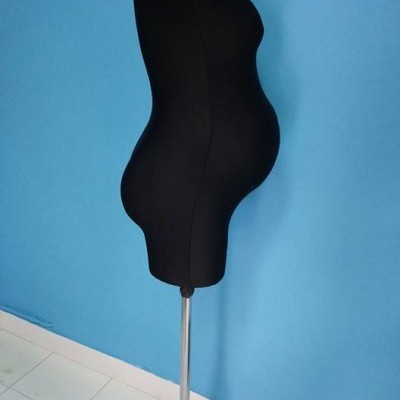 manequim preto com barriga saliente sobre suporte metálico num chão branco e parede azul