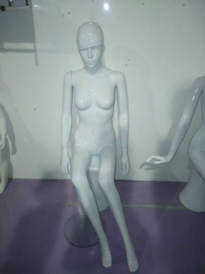 Mannequim feminino branco sentado, textura lisa e brilhante, sem roupas, em chão roxo.