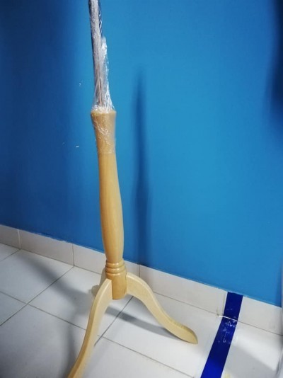 Suporte de manequim de madeira clara com base de três pernas, parcialmente envolvido em plástico, junto a uma parede azul