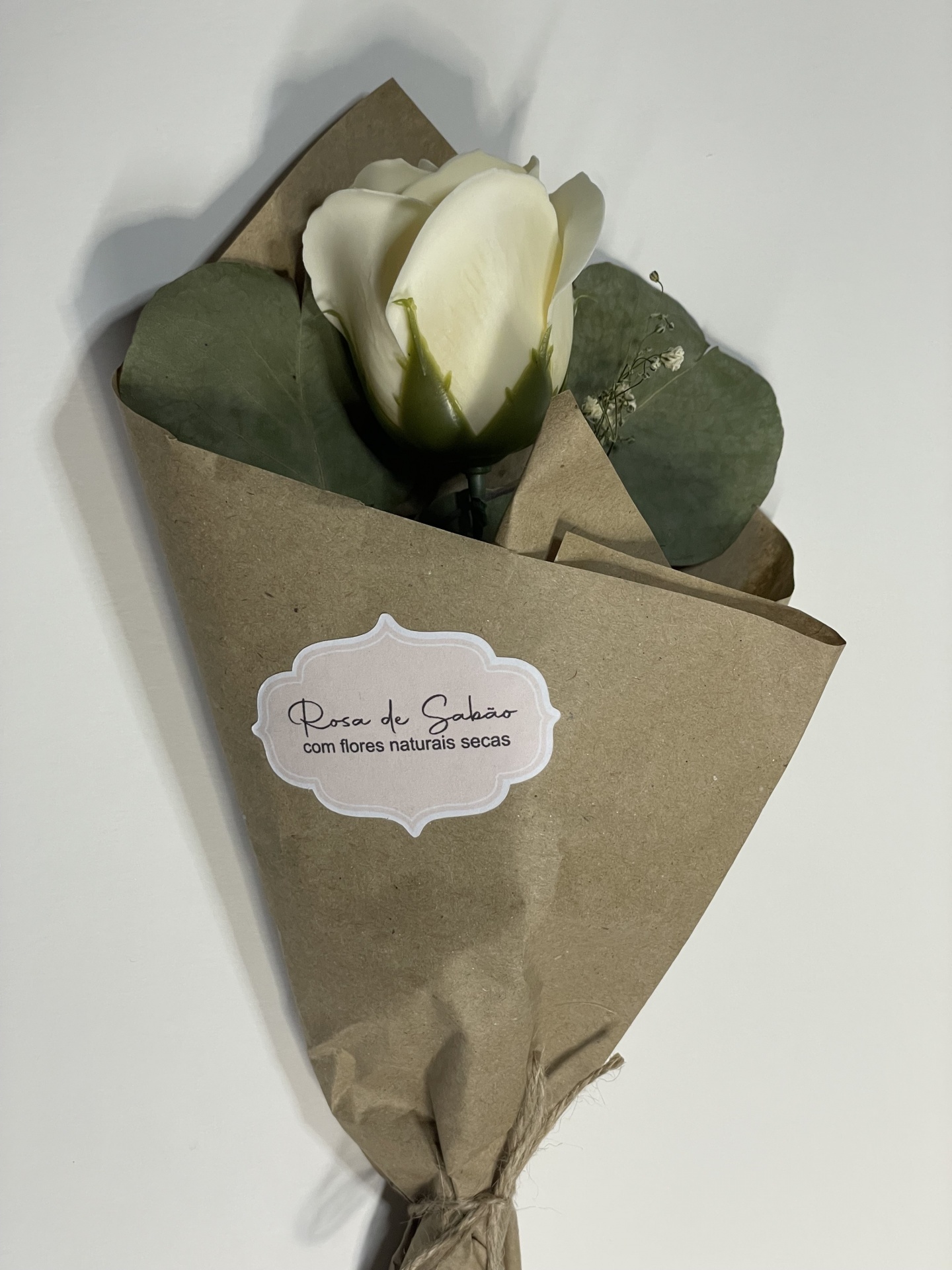 Ramo de rosa branca artificial com folhas verdes e flores naturais secas embrulhado em papel castanho