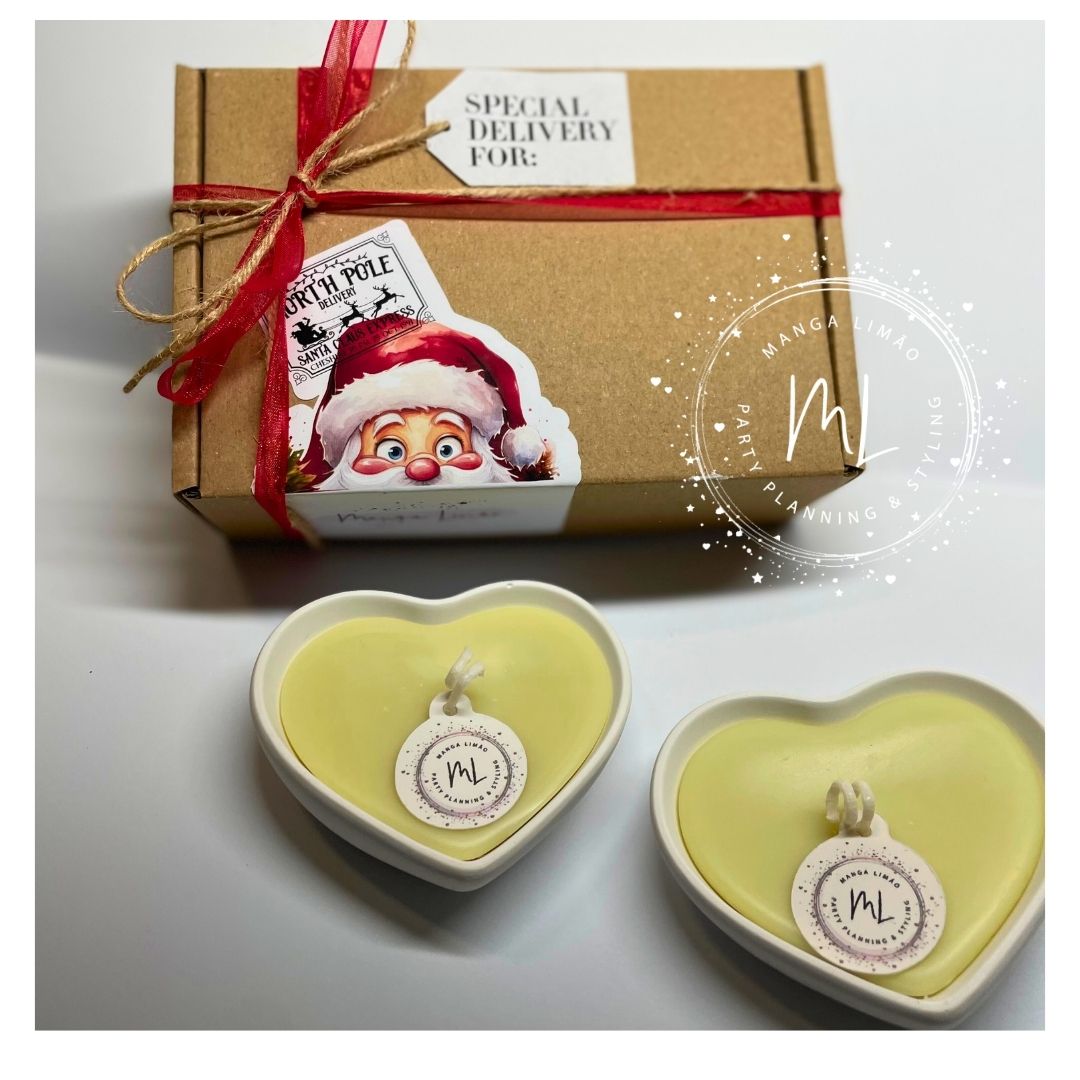 Duas velas amarelas em recipientes corações de cerâmica branca junto a caixa castanha com fita vermelha e etiqueta de Natal.