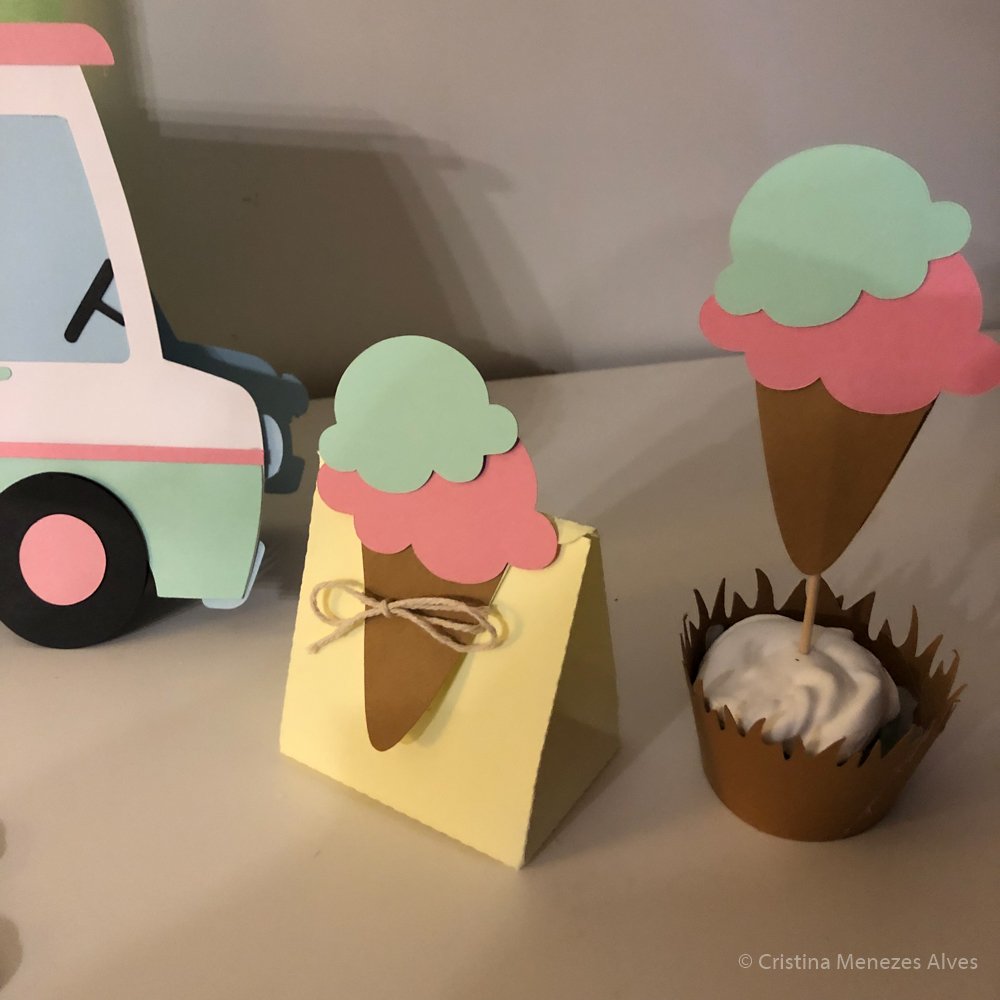 Decorações em papel de cones de gelado verdes e cor-de-rosa em caixa amarela com laço e base marrom com cobertura branca.